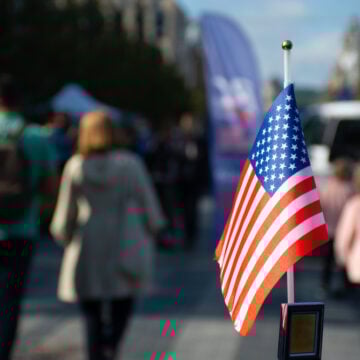American Flag Waving On A Car With People Walking In The Foreground