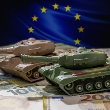 Toy Tanks Ride On Euro Banknotes Against The Background Of The NATO Flag