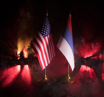 Closeup,,Flags,Of,Russia,And,Usa,Against,Destroyed,City,At Closeup Of Flags Of Russia And U.S.