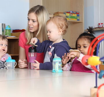 Kindergarten Or Nursery School Kids At A Table Drinking And Playing With Their Caregiver