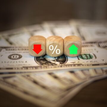 Three Dice, One With Red Arrow Pointing Down, Green Arrow Pointing Up, And Percentage Sign, Sitting On Stack Of Hundred Dollar Bills