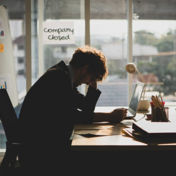 Person With Hand In Head At Desk With Company Closed Sign in Background
