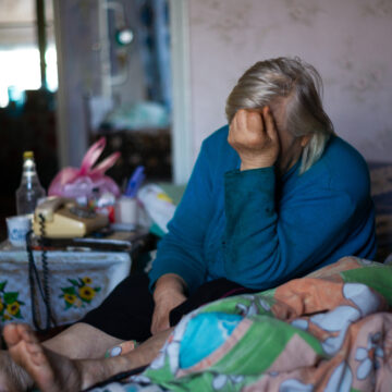 Sad, Old, Gray-Haired Woman Sitting On Her Bed