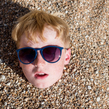 A Freckle-faced Boy With Sunglasses Up To His Neck In Rocky Sand