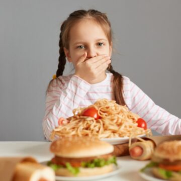 Little Girl With Braids Holds Her Hand Over Her Mouth While Sitting At A Table Filled With Food