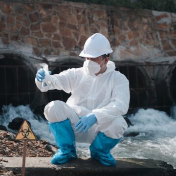 Ecologist In White Protection Suit Sampling Water From River With Test Tube For Toxic Chemicals