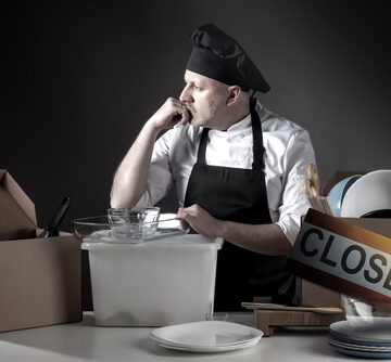 Restaurant Owner/Chef Overwhelmed By Financial Problems With Closing Sign And Kitchen Items Indicating Bankruptcy