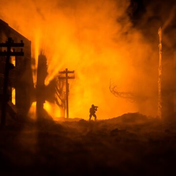 Army Sniper With Large Caliber Rifle Standing In The Middle Of Burning, Bombed Buildings