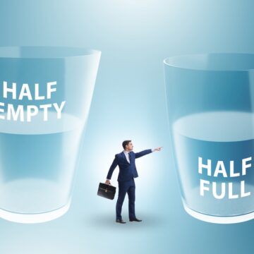 Illustration Of Businessman Between Two Glasses Of Water – One Half Empty And One Half Full