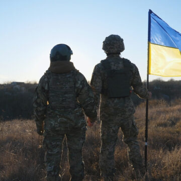 Two Ukrainian Soldiers With Flag Stand At The Edge Of A Battlefield