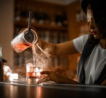 Woman,Bartender,In,A,White,T-shirt,And,Vest,Pours,A Female Bartender Pours A Mixed Drink