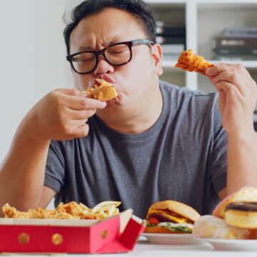 Overweight Man Gorging On A Large Pile Of Fast Food/Junk Food