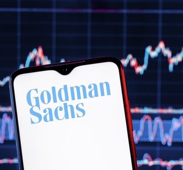 Smartphone With Goldman Sachs On Screen And Stock Graphs In The Background