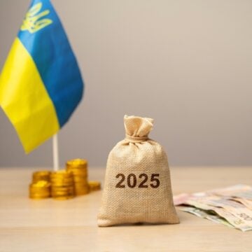 The Ukraine Flag With Stacks Of Gold Coins, Currency, And A Burlap Sack With The Year 2025 On The Front