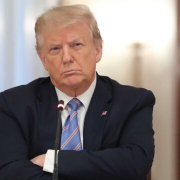 An Unsmiling Donald Trump Sits In Front Of A Microphone With His Arms Crossed