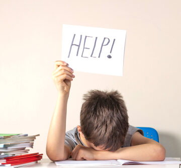 Tired And Frustrated Elementary School-Aged Boy Sitting At Table With His Classwork And Holding Up A Sign Reading "Help"