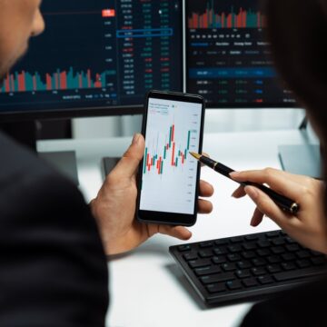 Two Stock Traders Reviewing Stock Prices On Mobile Phone And Computers