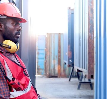 Portrait Of Young, Weary Worker Wearing Hard Hat And Headphones Leaning Against Shipping Container