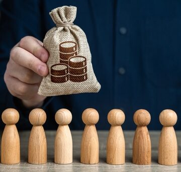 A Man's Hand Hold A Bag Of Coins Above, A Crowd Of Wooden People Pieces