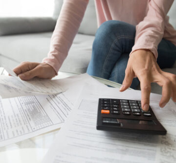 Woman's Hands Reaching For Calculator Atop A Pile Of Paperwork, Statements, Bills