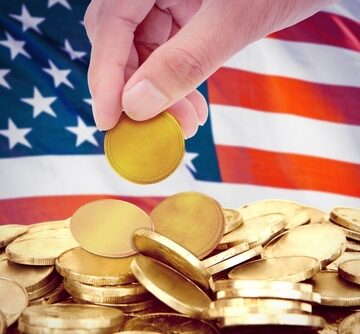Hand Dropping Gold Coin Atop Pile Of Gold Coins With An American Flag In Background