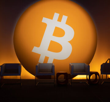 The Bitcoin Logo Against A Large Orange Circle Backdrop On A Stage With Chairs