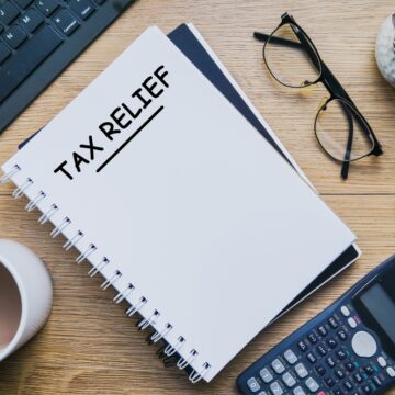 Financial Concept Image The Text "Tax Relief" On A Notepad, Calculator, Eyeglasses, Laptop Keyboard, And Cup Of Coffee