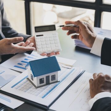 Realtor And Client With Calculator, Model House, Blueprints, And Financial Printouts