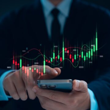 A Man's Hand Points To A Cell Phone With An Upward Stock Graph Superimposed Above The Phone