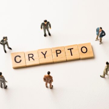 The Image Displays The Word "CRYPTO" Spelled Out With Wooden Letter Tiles, Surrounded By Miniature Figures Of People