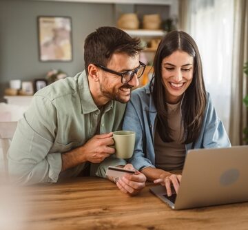 A Smiling Young Couple In Their Home Looking At A Laptop