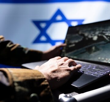 A Military Operative, Using A Laptop With The Israeli Flag In The Background