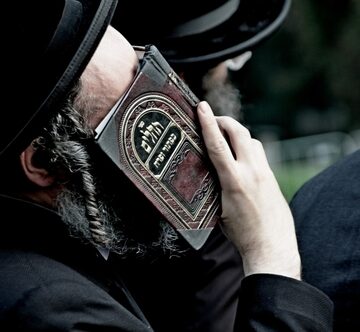 An Orthodox Jewish Man, Identifiable By His Black Hat And Peyot (Sidelocks), Holding A Religious Text To His Face, Likely In Prayer Or Study