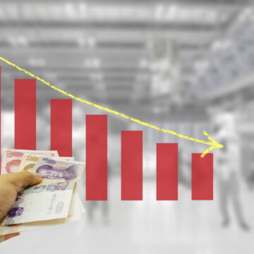 Concept Image With A Hand Holding A Stack Of Chinese Yuan Banknotes, A Bar Chart With Decreasing Red Bars, And A Downward-Pointing Yellow Arrow Concept Image With A Hand Holding A Stack Of Chinese Yuan Banknotes, A Bar Chart With Decreasing Red Bars, And A Downward-Pointing Yellow Arrow