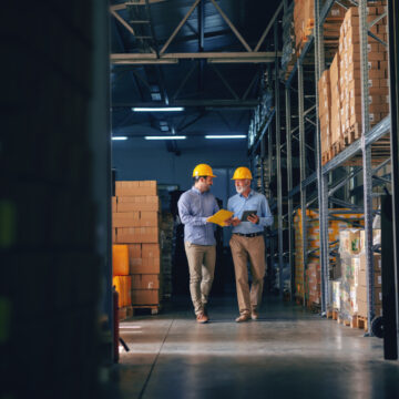 Two Men Wearing Yellow Hard Hats Walking Through A Warehouse