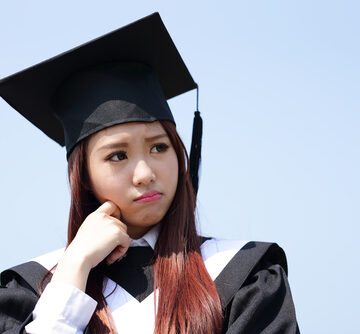 A Pensive College Graduate In Cap And Gown Against A Blue Sky