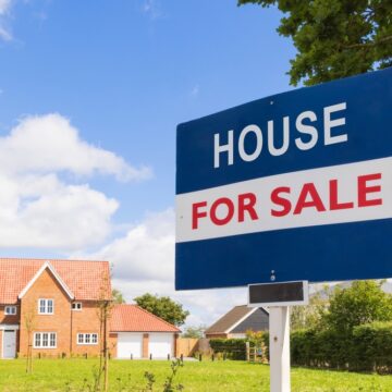 A Realtor "For Sale" Sign On Display Outside A Large Red, Brick Home A Realtor "For Sale" Sign On Display Outside A Large Red, Brick Home