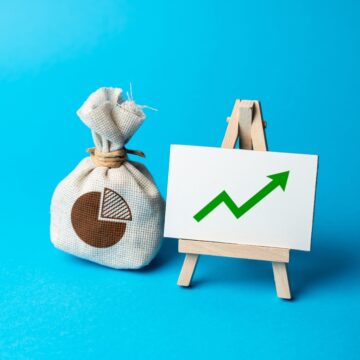 A Money Bag Next To A Board On A Easel With A Graph Showing A Green Up Arrow A Money Bag Next To A Board On A Easel With A Graph Showing A Green Up Arrow