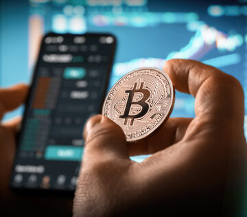 Concept Image Of A Day Trader Holding A Bitcoin, Mobile Phone With Data, And A Computer Display With Graphs In Background