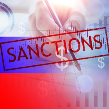 Concept Collage With Word "Sanctions" In Red, Currency Symbols, A Hand Holding A Pen, And Graphs In The Background