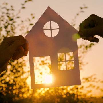 Photo Shows Two Hands Holding A Paper Cutout Of A House Against A Sunset Backdrop