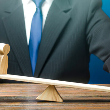 A Man Places One Green Wooden Figure On A Scale And It Tips The Balance Against Multiple Wooden Figures