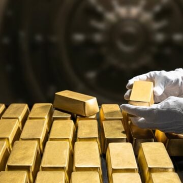 Banker's Hands In White Gloves Holding A Gold Bar From A Large Stack Of Gold Bars