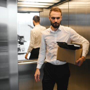 A Young Man Who Has Been Fired Or Is Leaving His Job, Carrying A Box Of His Personal Belongings In An Elevator A Young Man Who Has Been Fired Or Is Leaving His Job, Carrying A Box Of His Personal Belongings In An Elevator