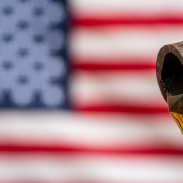 A Rusty Pipe With A Slow Drip Of Viscous, Amber Liquid, Likely Crude Oil, And A Blurred U.S. Flag In The Background