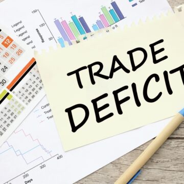Image Features A Yellow Sticky Note With The Words "TRADE DEFICIT" Written In Bold, Black Capital Letters, Placed On Top Of Several Financial Charts And A Calendar