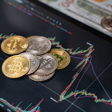 A Pile Of Bitcoin (BTC) Cryptocurrency Coins, Alongside U.S. Currency, Resting On A Screen Showing A Market Chart
