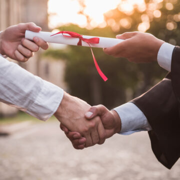 A graduate, Dressed In Academic Regalia, Receiving A Diploma And Shaking Hands With Another Person