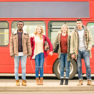 Four Young Friends Standing On A Sidewalk In Front Of A Traditional Red Double-decker Bus Famously Found In London, England