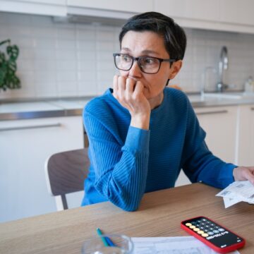 Stressed Mature Woman Surrounded By Bills, Receipts, And A Calculator At Her Kitchen Table